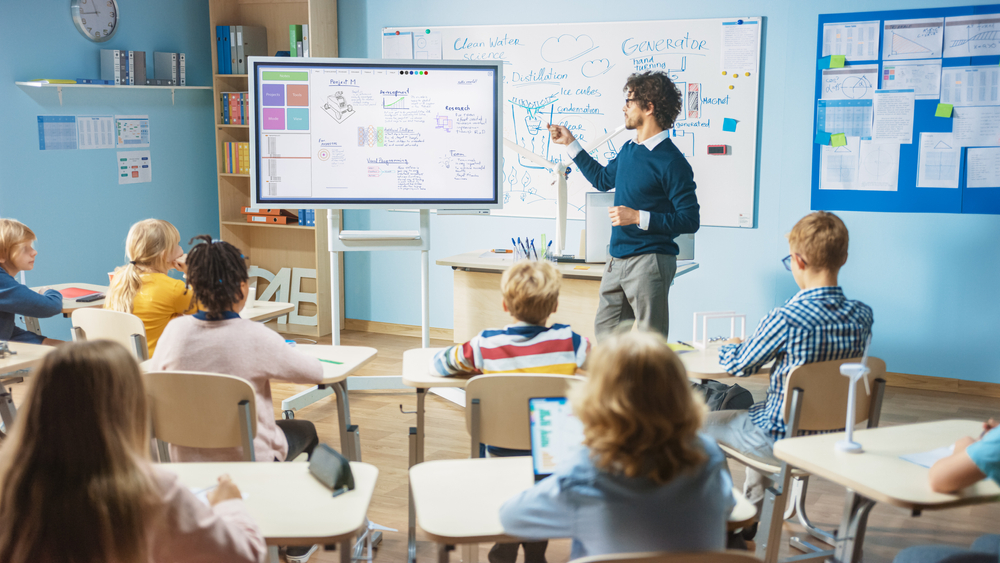 A imagem representa um professor apresentando uma aula interativa com auxílio de uma lousa digital em frente a uma turma de alunos atentos, representando um ambiente educacional moderno e dinâmico.